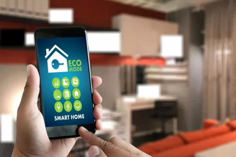 Im Vordergrund ist ein Smartphone mit einem Smart-Home-System zu sehen, das gerade von zwei Händen bedient wird. Im Hintergrund ist ein Wohnraum.